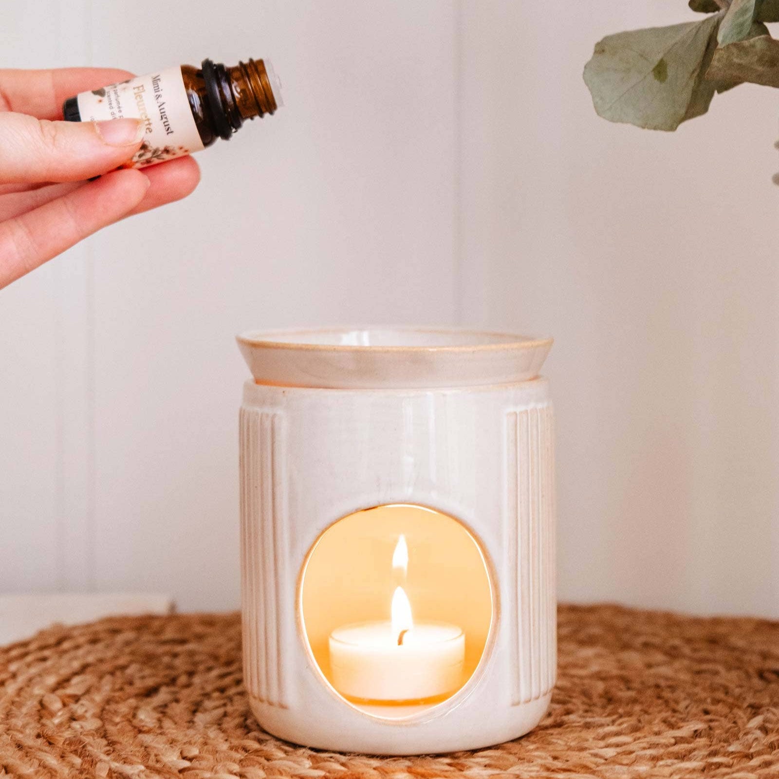 Person holding a bottle of essential oil above a lit candle in a ceramic holder with eucalyptus leaves in the background.