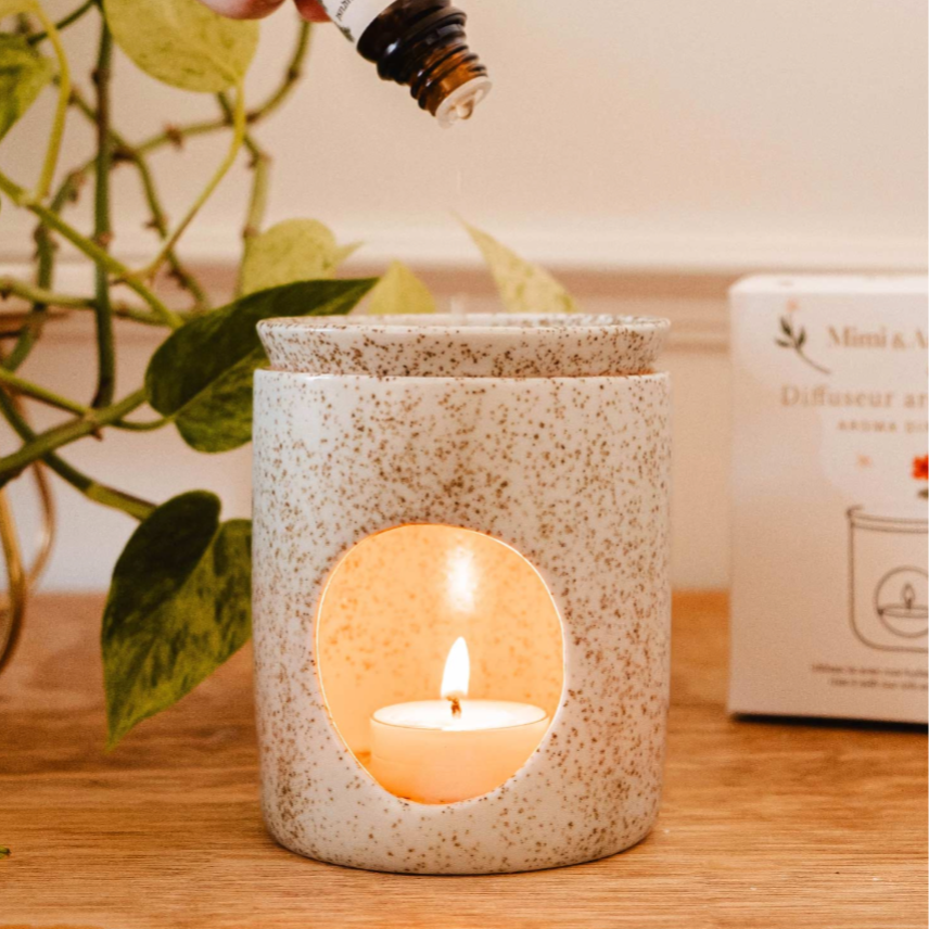Hand pouring essential oil into a diffuser with a candle and plant in the background