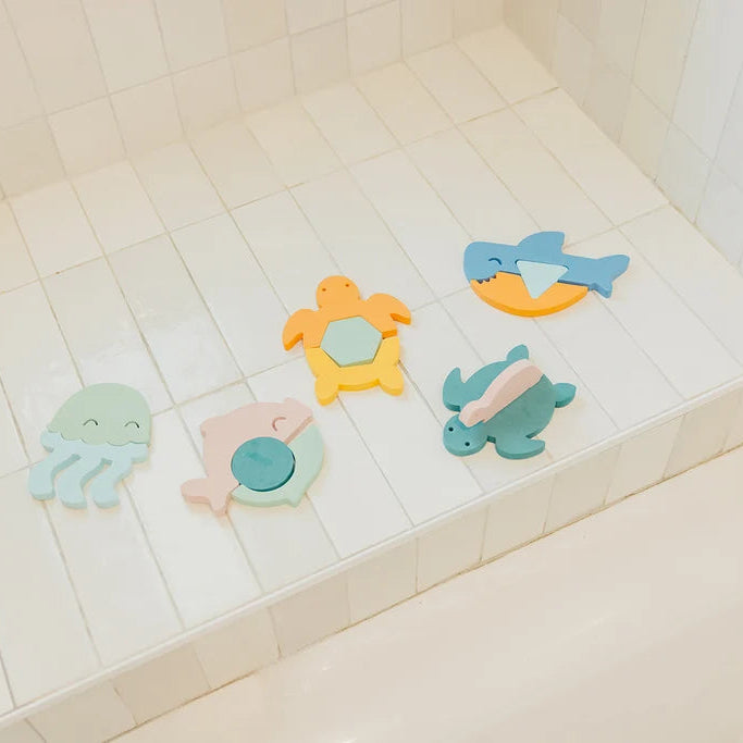 Four colorful animal-shaped bath toys arranged on a white tiled bathtub ledge. The toys include a light blue octopus, a pale pink fish, a yellow turtle, and a blue shark, all with soft pastel colors.