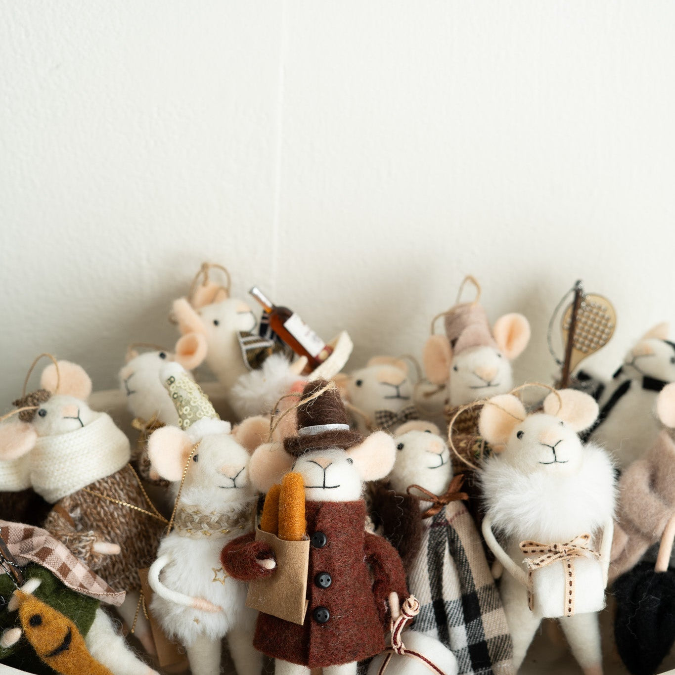 A group of small felted wool mouse ornaments dressed in various cozy outfits, including a brown coat with a hat and a plaid scarf, arranged in a white bowl against a plain white wall.