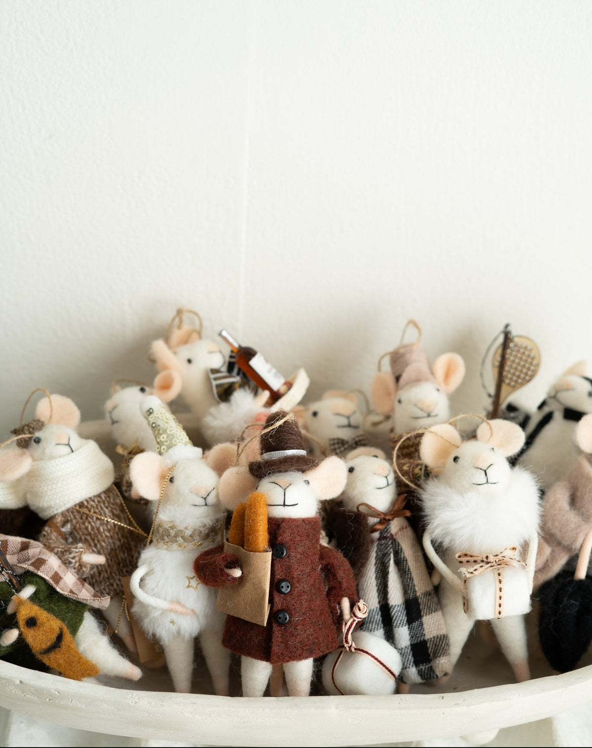 A group of small felted wool mouse ornaments dressed in various cozy outfits, including a brown coat with a hat and a plaid scarf, arranged in a white bowl against a plain white wall.