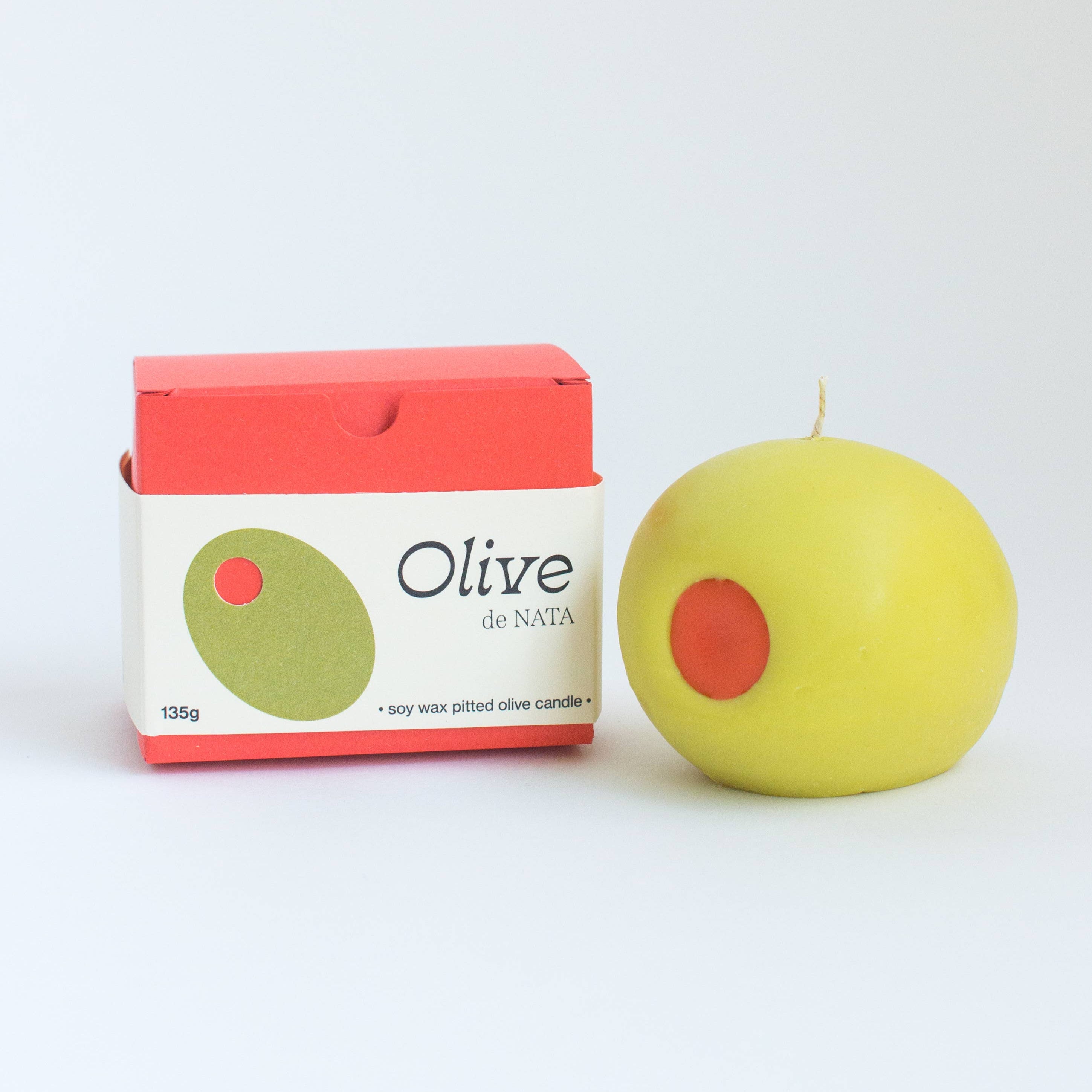 Yellow olive-shaped candle with a red center next to its packaging on a white background