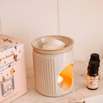Ceramic diffuser with a lit candle, packaging, and essential oil bottle on a shelf.