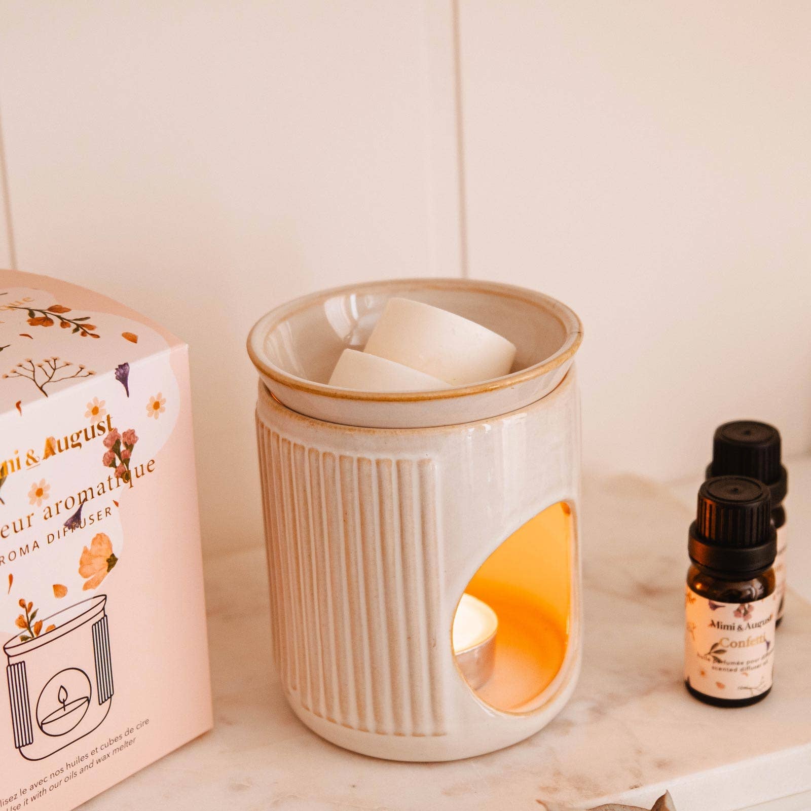 Ceramic diffuser with a lit candle, packaging, and essential oil bottle on a shelf.